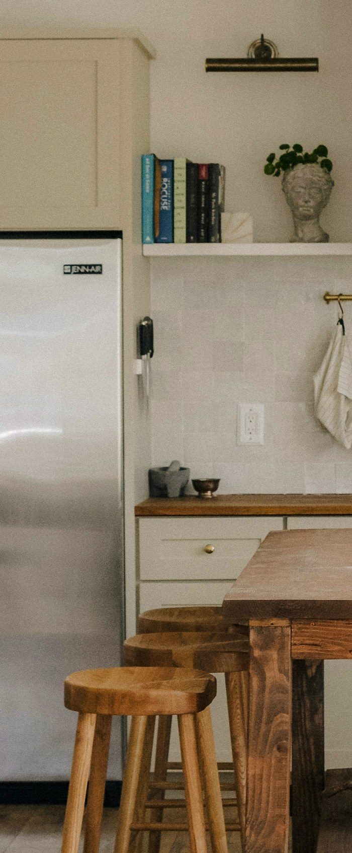 A stainless refrigerator in a warm kitchen, with cookbooks and a small plant on the open shelf above it.